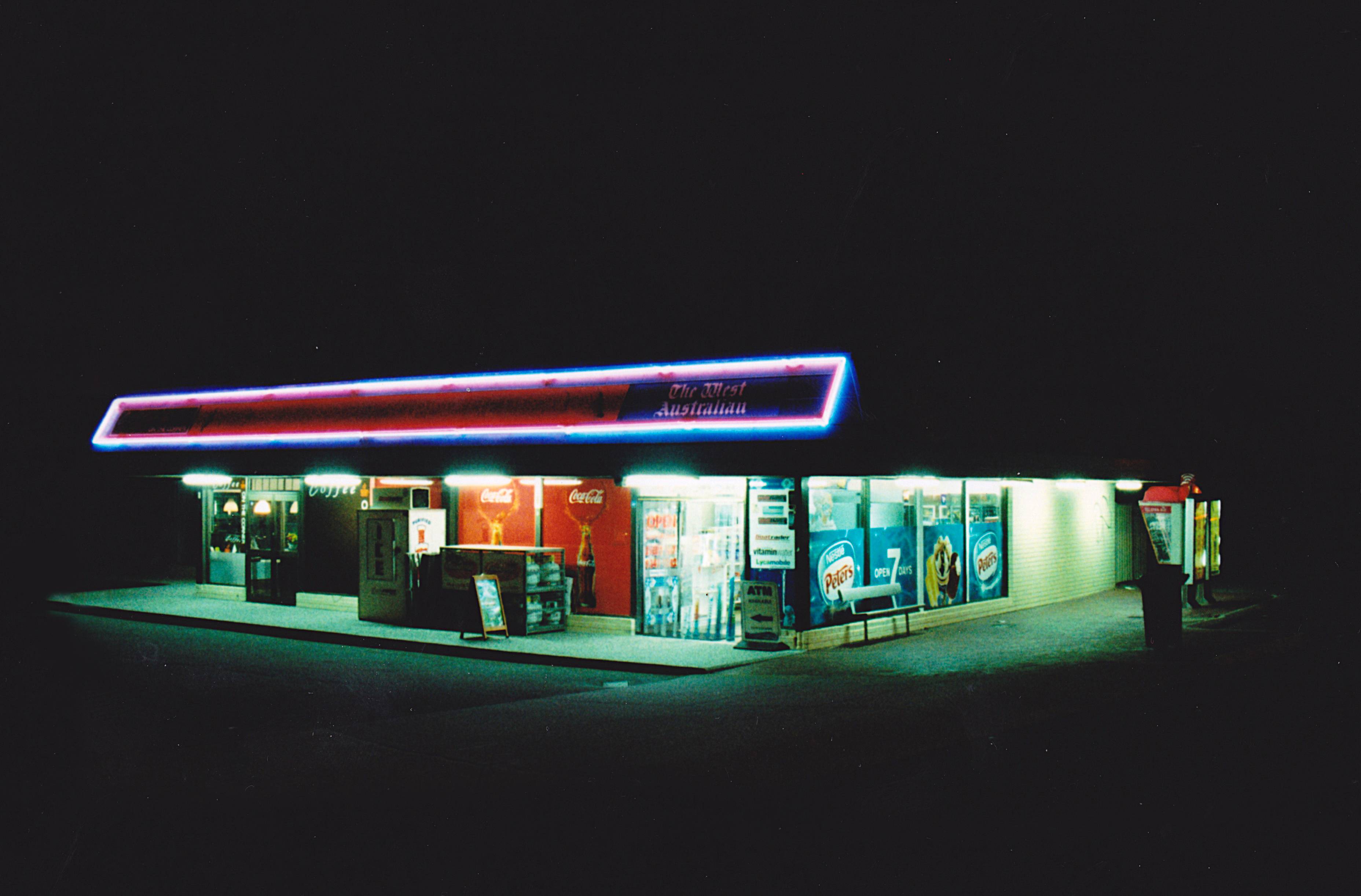 Okay, let's analyze this image using the requested theoretical frameworks. ## Visual Description The image presents a brightly lit, single-story convenience store, likely a gas station, at night. The exterior is predo...