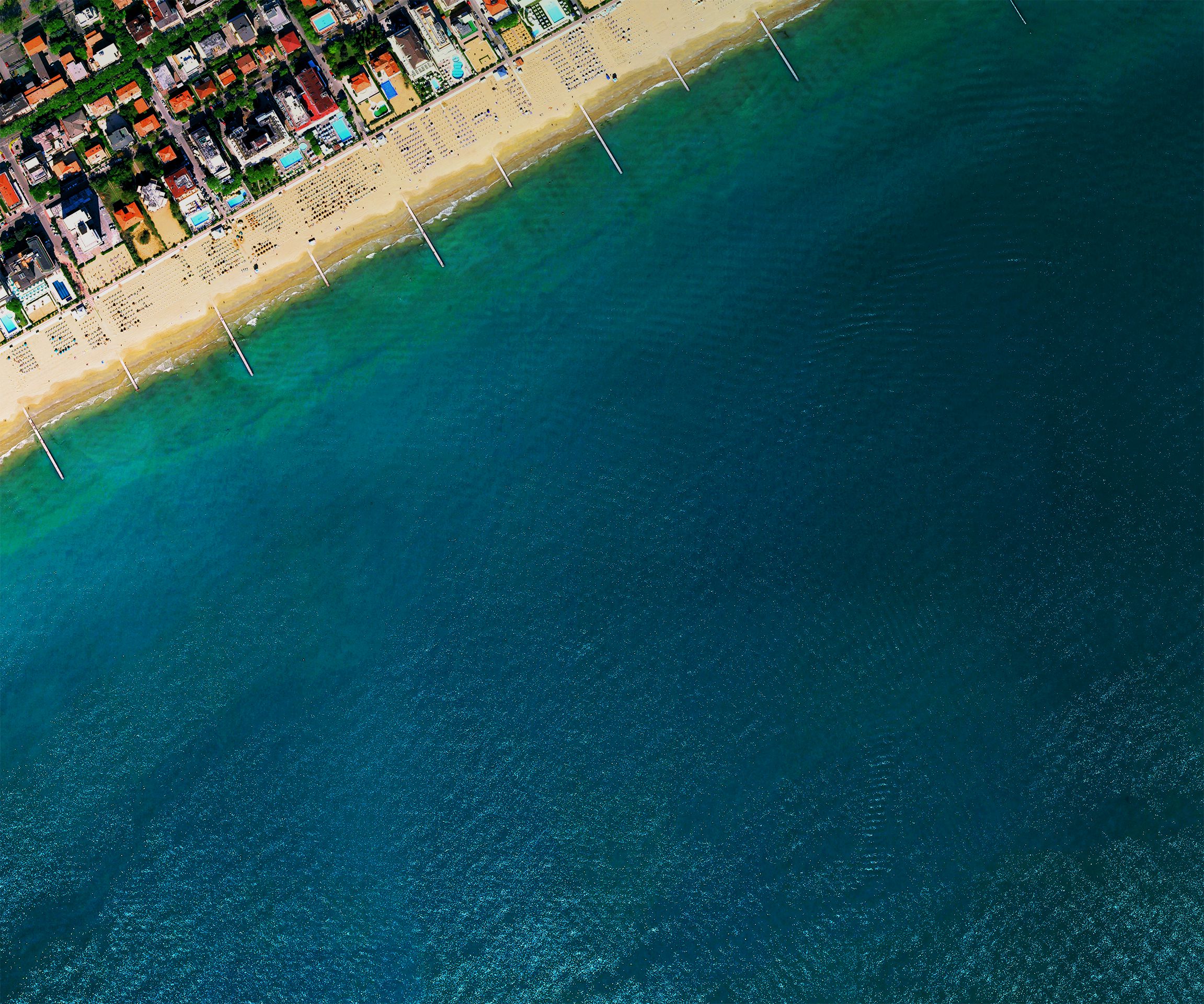 Okay, let's analyze the image through the requested lenses. **Visual Description** The image is a high-angle aerial photograph showing a coastline. Approximately 2/3 of the frame is dominated by the dark turquoise/dee...