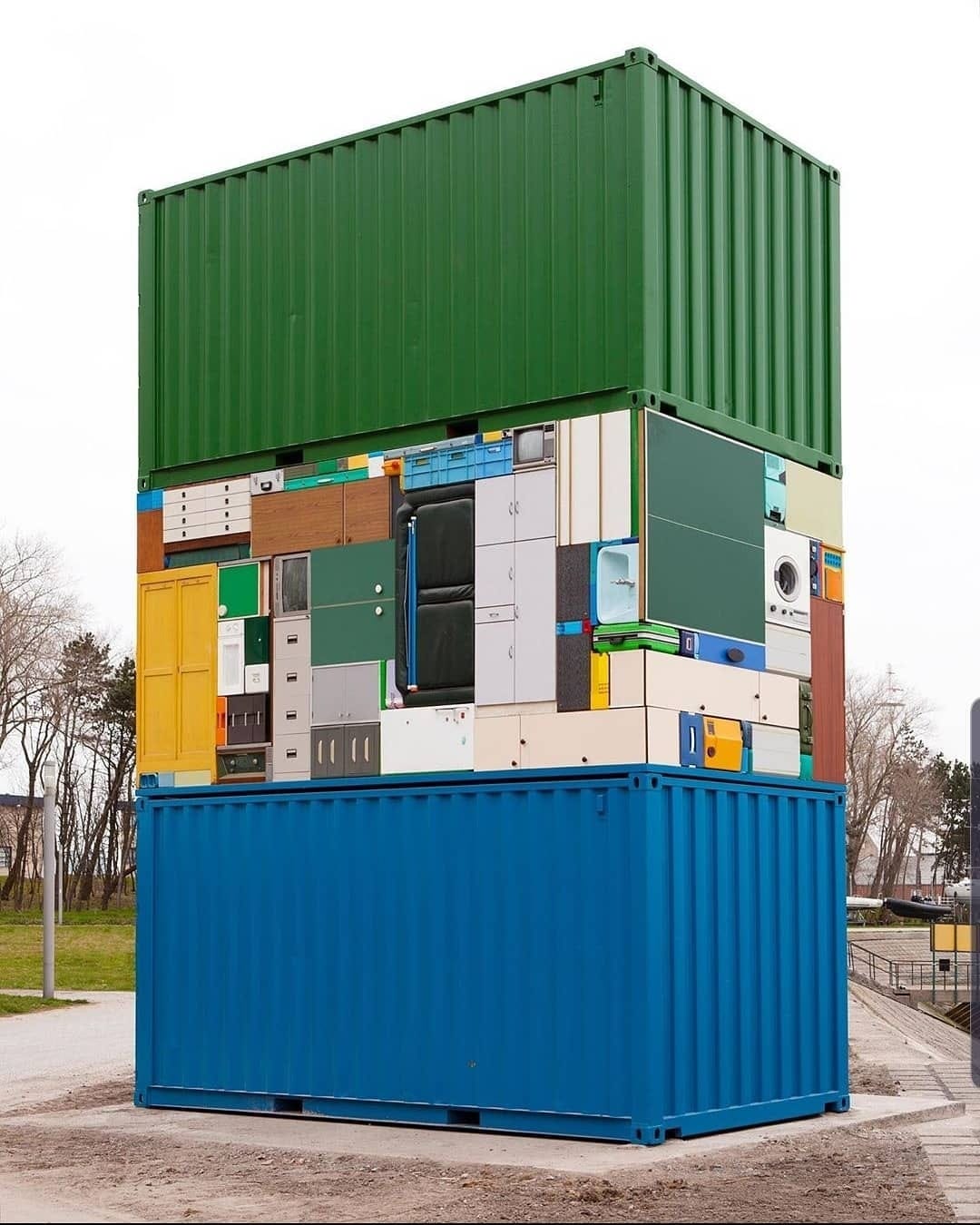 Okay, let's break down this striking image using the theoretical frameworks requested. **Visual Description** The image shows a tall, vertically stacked structure built from repurposed shipping containers. There are t...