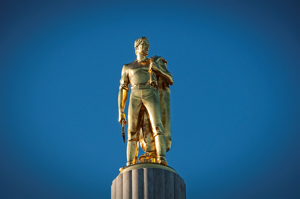 Okay, let's break down this image of a statue with several theoretical lenses. **Visual Description** The image depicts a bronze or gold-toned statue of a man, likely representing a historical figure, against a vibran...