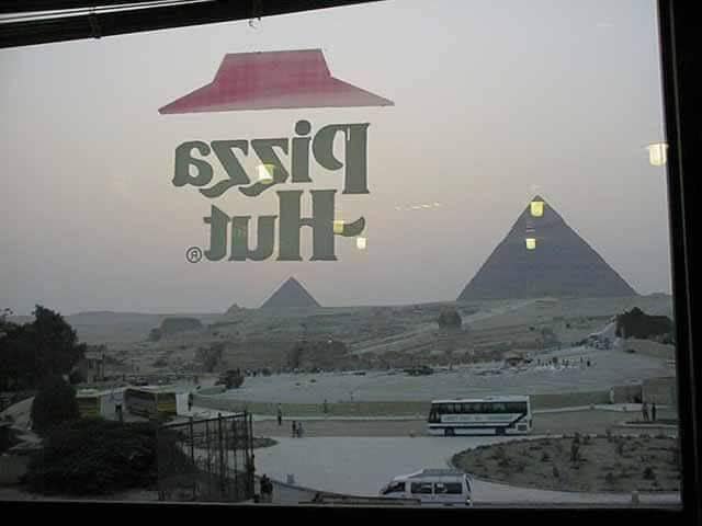 Okay, let's break down the image of the Pizza Hut sign superimposed over the Giza Pyramids through the lenses requested. ## Visual Description The photograph depicts a view through a glass window or storefront, likely...